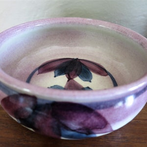 May include: A small, round ceramic bowl with a purple and blue floral design. The bowl has a light purple glaze and a white interior.