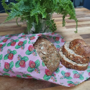May include: A pink floral patterned reusable food wrap with a loaf of multigrain bread inside. The wrap is made of beeswax, organic cotton, and tree resin.