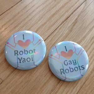 May include: Two circular pin-back buttons with a light blue holographic finish. The left button features a rainbow heart and the text I Robot Yaoi. The right button features a rainbow heart and the text I Gay Robots. Both have a line-art robot graphic.