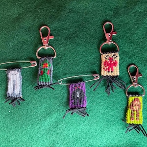 May include: Five woven textile charms featuring pixelated character designs. Two are attached to silver safety pins, and three are attached to silver lobster claw clasps. The designs include a white ghost, a green figure, a purple monster, a beige creature, and a yellow figure.