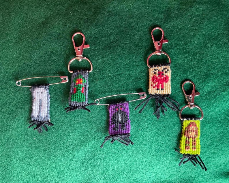 Puede incluir: Cinco dijes textiles tejidos con dise&ntilde;os de personajes pixelados. Dos est&aacute;n sujetos a imperdibles plateados y tres a cierres de mosquet&oacute;n plateados. Los dise&ntilde;os incluyen un fantasma blanco, una figura verde, un monstruo morado, una criatura beige y una figura amarilla.
