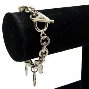 Könnte beinhalten: Ein silberfarbenes Gliederarmband mit Knebelverschluss. Das Armband enthält mehrere kleine, runde Anhänger, die an den Gliedern befestigt sind, wobei ein Anhänger ein sichtbares, eingraviertes Design in Form eines geschriebenen Buchstabens aufweist.