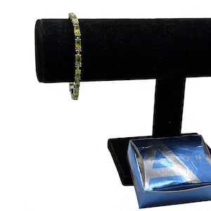 May include: A silver-toned link bracelet featuring a series of oval-shaped light green peridot gemstones alternating with small clear stones. The bracelet is displayed on a black T-bar stand next to a small blue box with the letter A visible on top.
