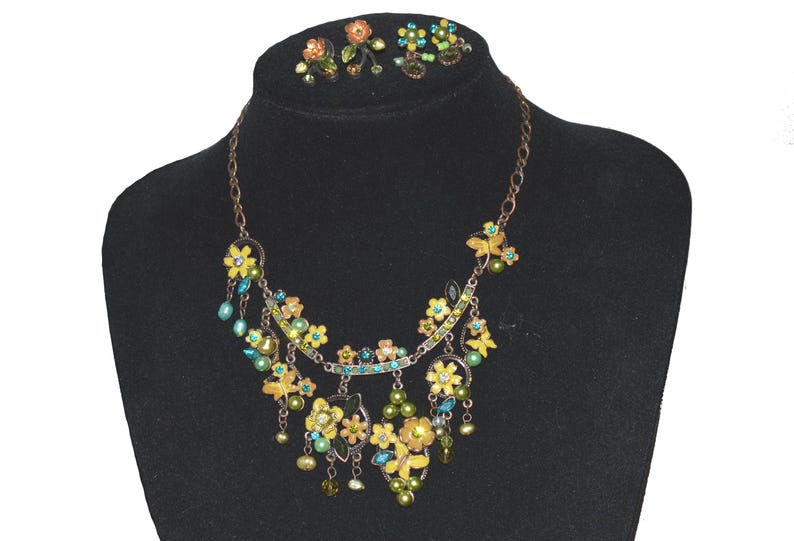 May include: A floral necklace and matching earrings set. The necklace features yellow and green flower pendants with blue and green accents, and dangling beads. The earrings have similar floral designs. The jewelry is displayed on a black mannequin.