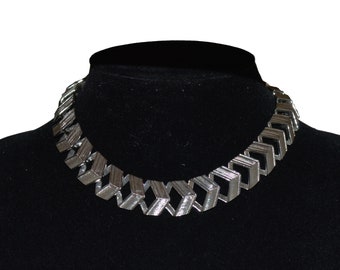 May include: A silver-toned necklace with a geometric design. The necklace features a series of interconnected, three-dimensional square shapes. The necklace is displayed on a black mannequin, showcasing its design and texture.