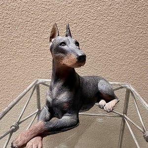 May include: A Doberman Pinscher figurine featuring a textured black and grey coat with tan markings on the muzzle and paws. The dog is depicted in a lying position with its front legs crossed, showcasing pointed ears and a focused expression.