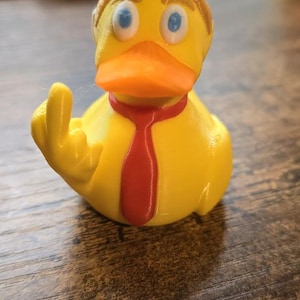 May include: A yellow rubber duck with a red tie and brown hair, giving the middle finger.