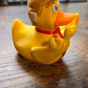 May include: A yellow rubber duck with a red tie and a blonde hairpiece. The duck is making an OK sign with its right wing.