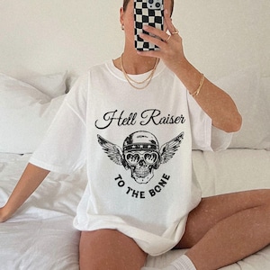 May include: White oversized t-shirt with a black graphic of a skull wearing a helmet and wings. The text "Hell Raiser to the Bone" is printed below the skull.