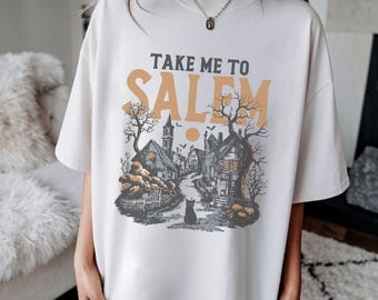 May include: Cream-colored t-shirt with the text "TAKE ME TO SALEM" in orange and gray. The graphic depicts a spooky town scene with houses, trees, and a black cat. The shirt has a relaxed fit.