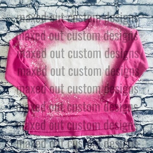 May include: A bright pink long-sleeve sweatshirt featuring a large, circular bleached white area in the center. The garment has a crew neckline, ribbed cuffs, and a straight hemline, with white speckled bleach patterns across the fabric surface.