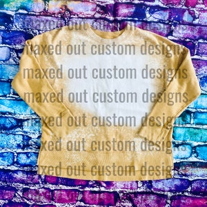 May include: A mustard yellow long-sleeve sweatshirt featuring a large, central bleached white area with a distressed, speckled effect. The garment has a crew neckline, ribbed cuffs, and a straight hemline. The text maxed out custom designs is overlaid.