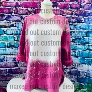 May include: A long-sleeve crewneck sweatshirt in a vibrant hot pink color featuring a large, circular bleached white area in the center. The garment has a distressed, splattered bleach effect throughout the fabric and a straight hemline.
