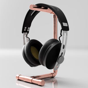 Pode incluir: Um suporte para fones de ouvido de cobre com um par de fones de ouvido pretos apoiados nele. O suporte é feito de tubo de cobre e tem um design simples e moderno.