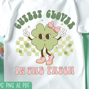 Könnte beinhalten: Weißes T-Shirt mit einer Cartoon-Figur eines vierblättrigen Kleeblatts mit rosa Schleife und Stiefeln, die Friedenszeichen zeigt. Der Text lautet "Cutest Clover in the Patch" in grüner und rosa Retro-Schrift, mit kariertem Hintergrund.