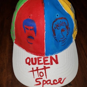 Può includere: Un cappello da baseball bianco con una corona rossa, blu, verde e gialla. La corona ha due volti dipinti su di essa. Il visore del cappello è bianco con le parole "QUEEN Hot Space" scritte in vernice rossa.