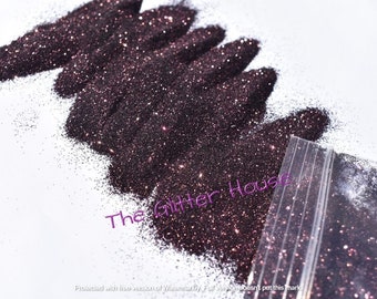 May include: Dark brown glitter is scattered across a white surface, with some contained in a clear plastic bag. The glitter has a sparkling effect, catching the light. The text "The Glitter House" is visible in the image.
