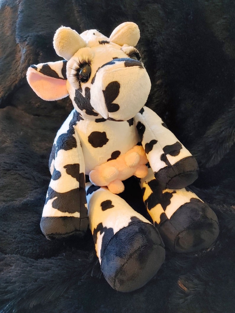 May include: A plush cow toy with black and white spots, large eyelashes, and a pink udder. The cow has black hooves and is sitting on a dark, textured surface. The ears are light tan with pink insides.
