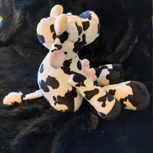 May include: A plush cow toy in black and white spots, with pink accents on the tongue and udders. The stuffed animal has floppy ears and is lying on a black, textured surface. A cute and cuddly farm animal.