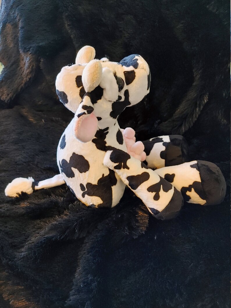May include: A plush cow toy in black and white spots, with pink accents on the tongue and udders. The stuffed animal has floppy ears and is lying on a black, textured surface. A cute and cuddly farm animal.