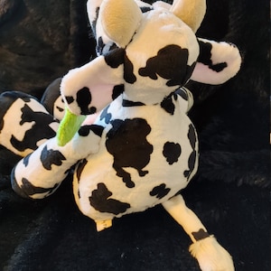 May include: A plush cow toy with a black and white spotted pattern. The toy has beige ears, a pink inner ear, and a green and pink object in its mouth. The cow is lying on a black, textured surface.