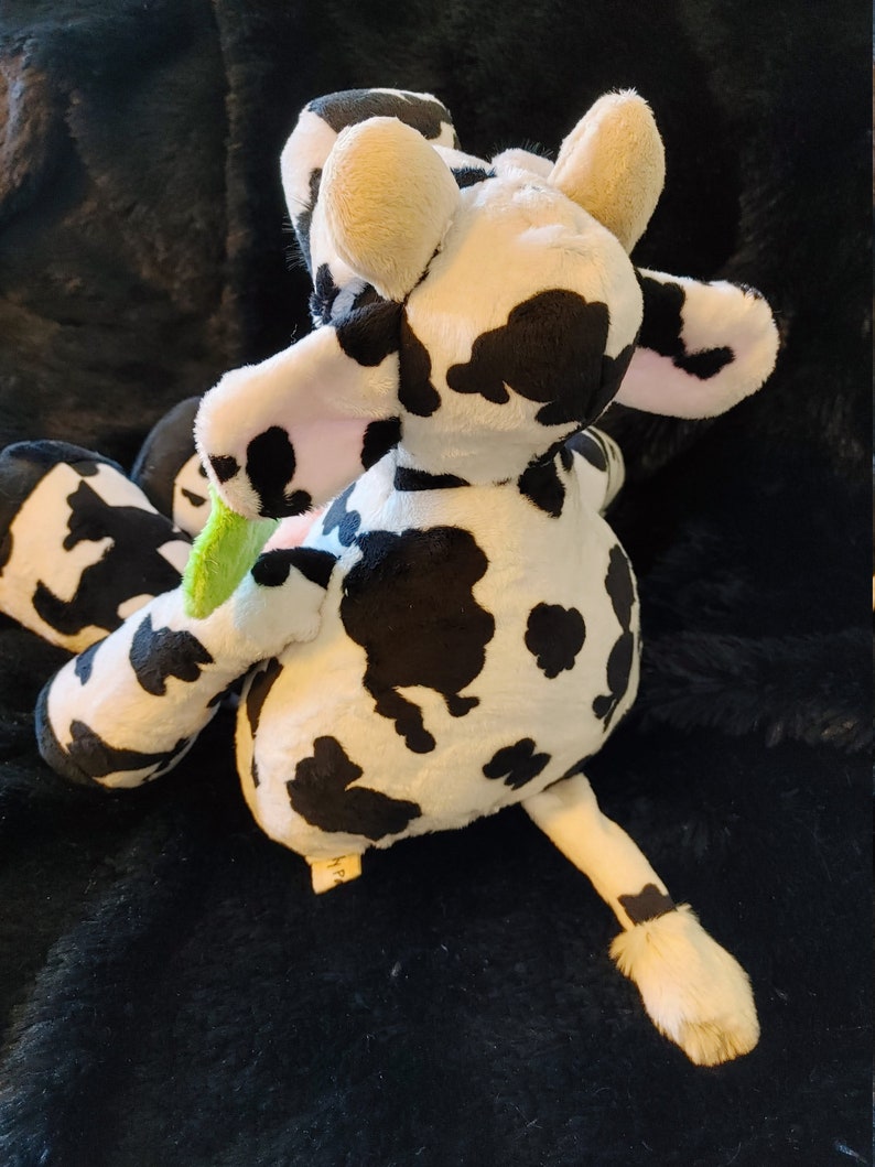 May include: A plush cow toy with a black and white spotted pattern. The toy has beige ears, a pink inner ear, and a green and pink object in its mouth. The cow is lying on a black, textured surface.