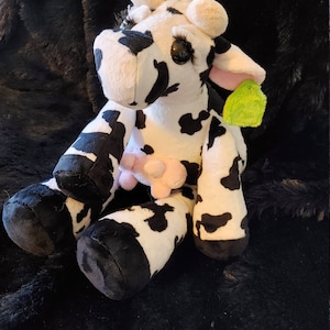 May include: A plush cow toy with black and white spots, large eyelashes, and pink ears. The stuffed animal has a pink udder and a green tag attached. The cow is sitting on a black, textured surface.
