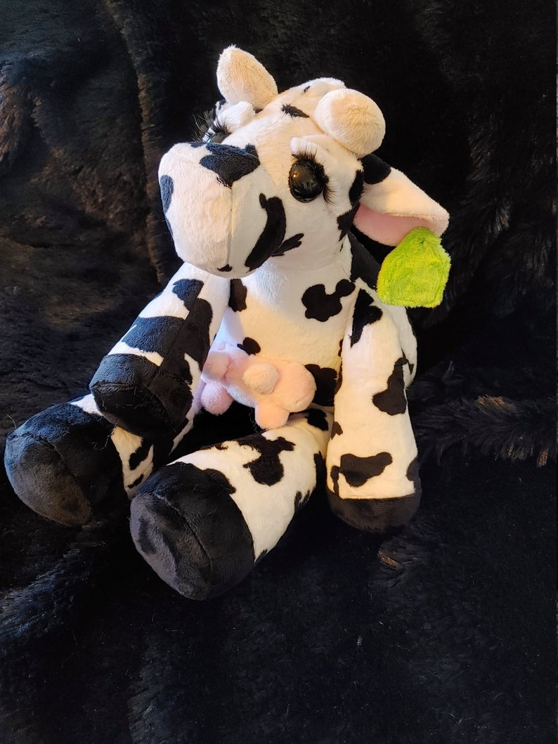 May include: A plush cow toy with black and white spots, large eyelashes, and pink ears. The stuffed animal has a pink udder and a green tag attached. The cow is sitting on a black, textured surface.
