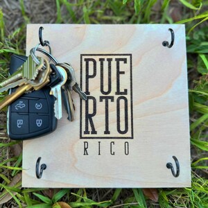 K&ouml;nnte beinhalten: Ein h&ouml;lzerner Schl&uuml;sselhalter mit dem Text "PUERTO RICO" in einem schwarzen rechteckigen Rahmen. Der Schl&uuml;sselhalter hat vier schwarze Haken und einen Satz Schl&uuml;ssel, einschlie&szlig;lich eines Autoschl&uuml;ssels.