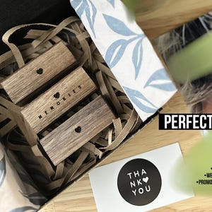 Puede incluir: Tres bloques de madera apilados uno encima del otro en una caja. Los bloques son marrones y tienen un coraz&oacute;n y la palabra "MEMORIES" grabada en ellos. La caja est&aacute; forrada con papel marr&oacute;n y tiene un papel estampado blanco y azul en el lateral. Una tarjeta con el texto "THANK YOU" es visible en la esquina inferior derecha de la imagen.