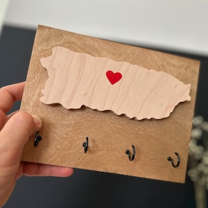 May include: Wooden key holder featuring a cutout of the island of Puerto Rico with a red heart in the centre. The key holder has four hooks for hanging keys.