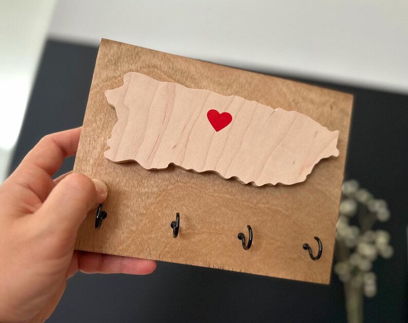 May include: Wooden key holder featuring a cutout of the island of Puerto Rico with a red heart in the centre. The key holder has four hooks for hanging keys.