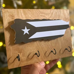 K&ouml;nnte beinhalten: Schl&uuml;sselhalter aus Holz mit der Umrissform von Puerto Rico mit dem Flaggen-Design in Schwarz, Wei&szlig; und einem Stern. Der Schl&uuml;sselhalter hat vier schwarze Metallhaken und ist aus hellbraunem Holz gefertigt.