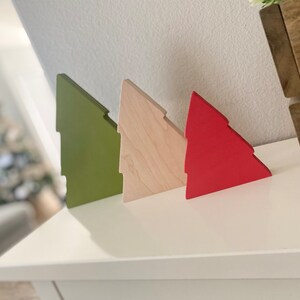 May include: Three wooden Christmas tree ornaments in different colors: green, natural wood, and red. The ornaments are arranged in a row on a white surface.