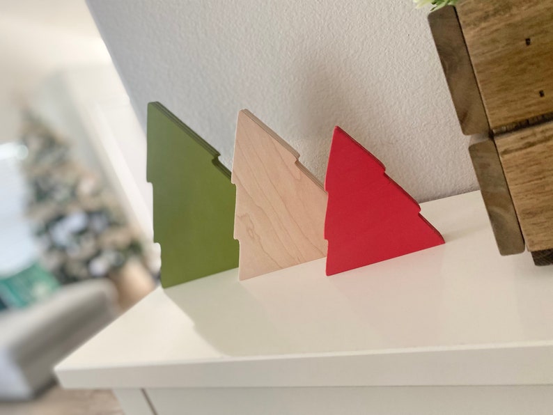 May include: Three wooden Christmas tree ornaments in different colors: green, natural wood, and red. The ornaments are arranged in a row on a white surface.