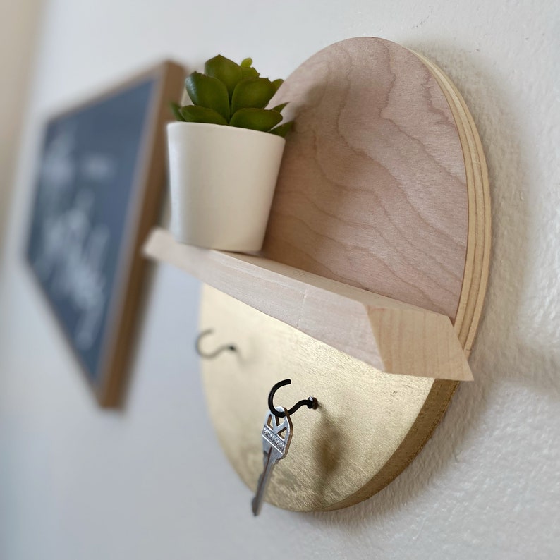 K&ouml;nnte beinhalten: Ein Schl&uuml;sselhalter aus Holz mit einem Regal und zwei Haken. Das Regal ist goldfarben lackiert und hat eine kleine Topfpflanze darauf. Der Schl&uuml;sselhalter ist an einer wei&szlig;en Wand montiert.