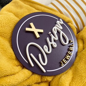 May include: A purple round wooden sign with a white outline and the words "+ Design Jewelry" in white and gold lettering.