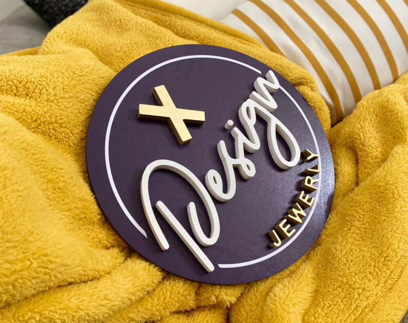 May include: A purple round wooden sign with a white outline and the words "+ Design Jewelry" in white and gold lettering.