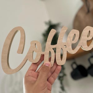 Pu&ograve; includere: Un cartello di legno con la scritta "Coffee" in corsivo. Il cartello &egrave; in legno chiaro ed &egrave; tenuto in mano. Lo sfondo &egrave; sfocato, ma include una mensola e delle tazze nere.