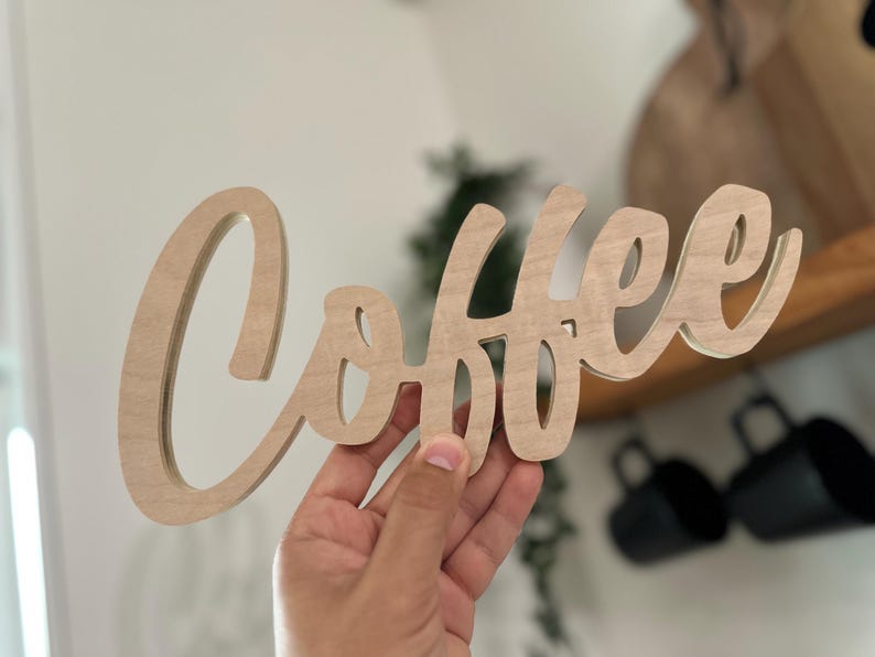 Pu&ograve; includere: Un cartello di legno con la scritta "Coffee" in corsivo. Il cartello &egrave; in legno chiaro ed &egrave; tenuto in mano. Lo sfondo &egrave; sfocato, ma include una mensola e delle tazze nere.