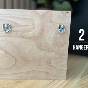 K&ouml;nnte beinhalten: Ein helles Holzquadrat mit zwei Metallaufh&auml;ngern. Der Text "2 HANGERS" ist wei&szlig; auf der rechten Seite des Holzes aufgedruckt. Die Holzmaserung ist sichtbar.