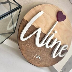 May include: A round wooden sign with a light brown and dark brown wood grain pattern. The word "wine" is written in white raised letters on the sign. A small red heart is attached to the top of the sign. A small white graphic of two wine glasses clinking is in the bottom center of the sign.