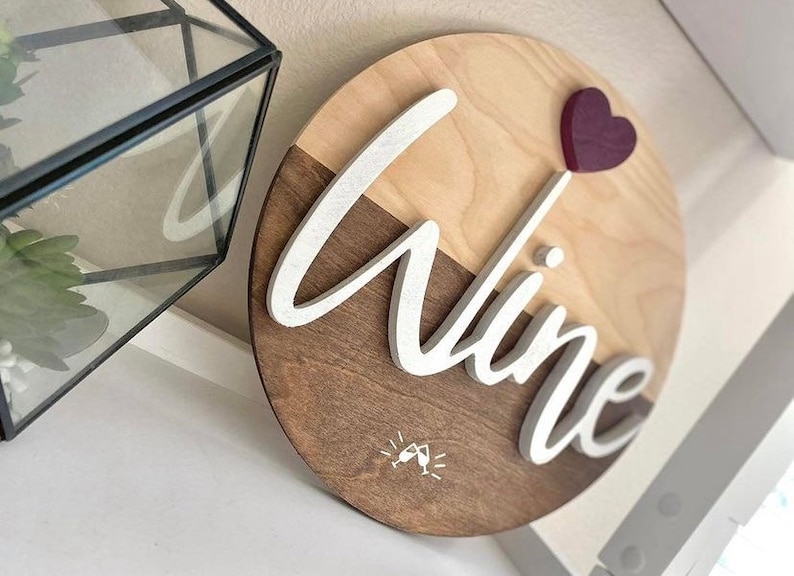 May include: A round wooden sign with a light brown and dark brown wood grain pattern. The word "wine" is written in white raised letters on the sign. A small red heart is attached to the top of the sign. A small white graphic of two wine glasses clinking is in the bottom center of the sign.