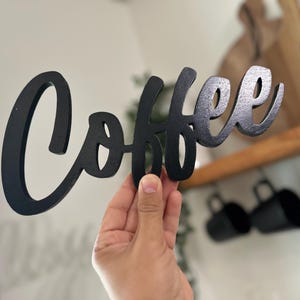May include: A black wooden sign in a cursive font that spells out the word "Coffee". The sign is held up in front of a blurred background of a kitchen with mugs and wooden cutting boards.