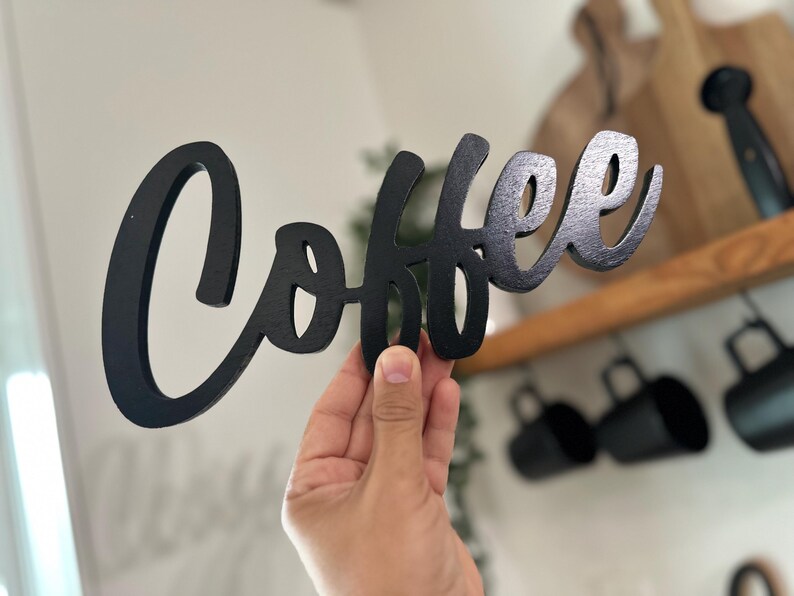 May include: A black wooden sign in a cursive font that spells out the word "Coffee". The sign is held up in front of a blurred background of a kitchen with mugs and wooden cutting boards.
