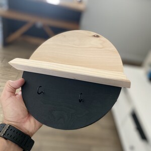 K&ouml;nnte beinhalten: Ein Schl&uuml;sselhalter aus Holz mit einem schwarz lackierten Halbkreis und zwei Haken. Die obere H&auml;lfte des Schl&uuml;sselhalters ist aus Naturholz.