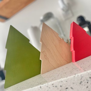 May include: Three wooden Christmas tree ornaments in different colors: green, natural wood, and red. The ornaments are arranged in a row on a white countertop.
