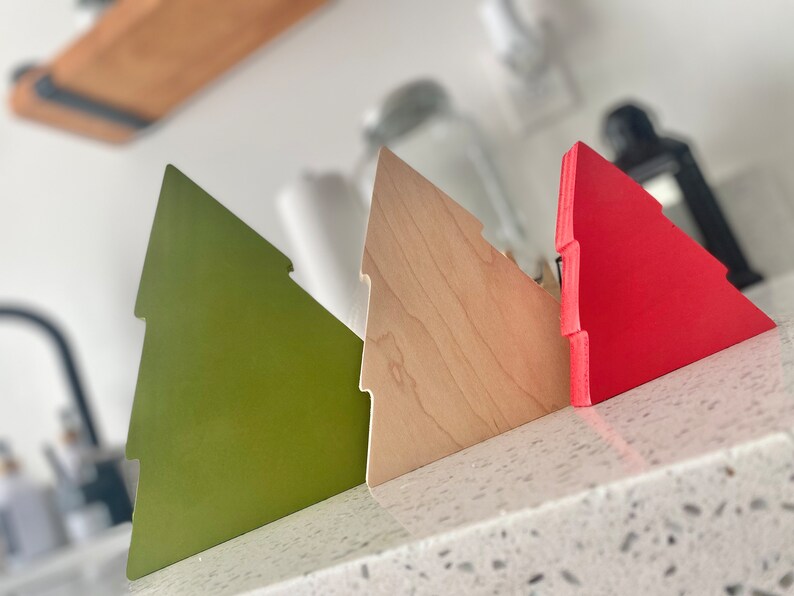 May include: Three wooden Christmas tree ornaments in different colors: green, natural wood, and red. The ornaments are arranged in a row on a white countertop.