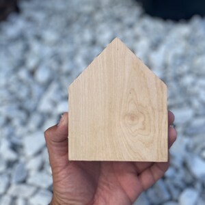 May include: A plain, unfinished wooden house shape, held in a hand, against a background of white gravel.