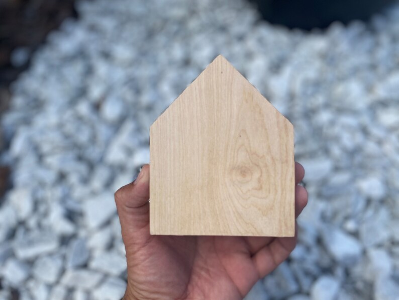 May include: A plain, unfinished wooden house shape, held in a hand, against a background of white gravel.